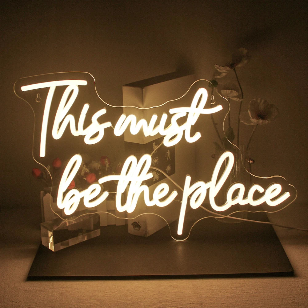 L'image montre un panneau néon LED vibrant sur lequel est écrit « This must be the place » en écriture cursive.