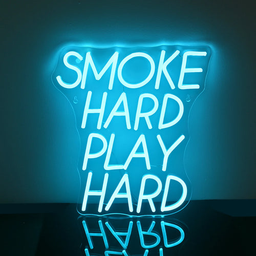 Na zdjęciu widoczny jest jaskrawy neon LED, na którym widnieje pogrubiony, drukowany napis „Smoke Hard Play Hard”.