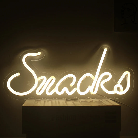 The image showcases a vibrant LED neon sign in the shape of the word "Snacks" in a playful, cursive font.