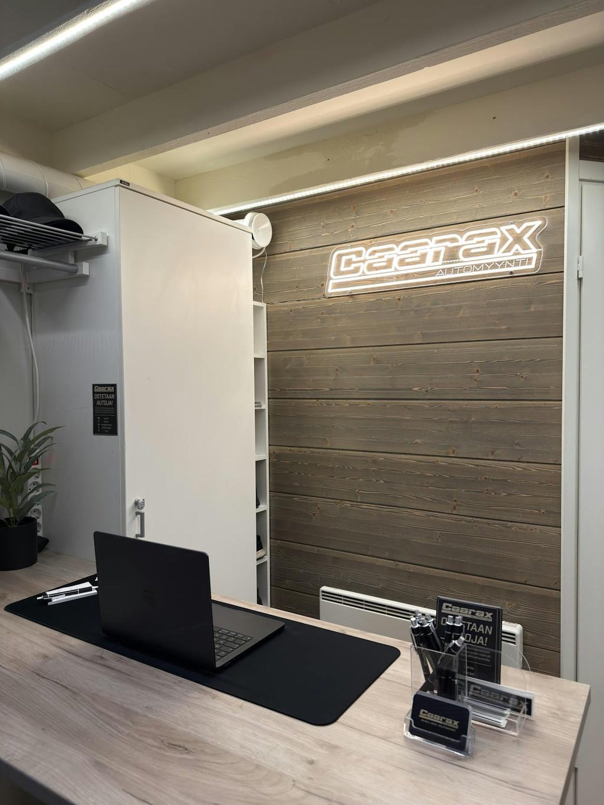 Logo Caarax Automyynti z teple bílého LED neonu instalované na kancelářské stěně s dřevěným obložením.